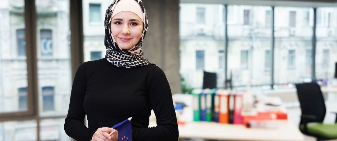 Junge Frau mit Kopftuch steht in einem Büro mit einer Europaflagge in der Hand