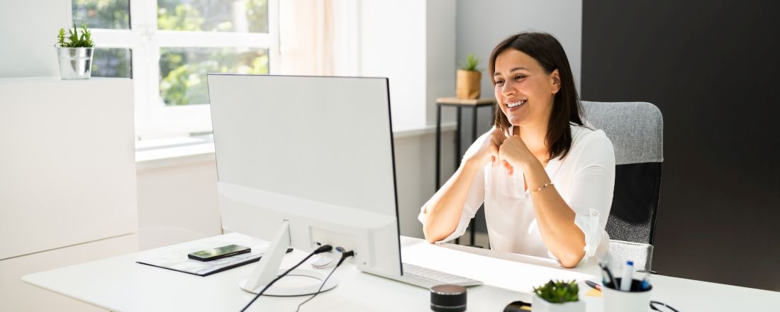 Führungskraft sitzt in ihrem Büro vor einem großen Bildschirm. Ihr Büro ist hell und geräumig. Sie trägt eine weiße Bluse und schulterlanges braunes Haar.