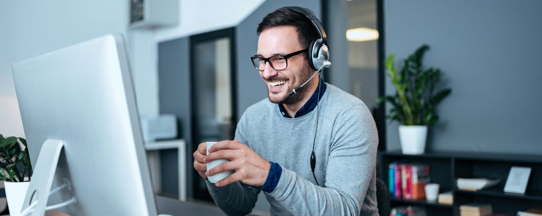 Mann in einem grauen Pullover sitzt in seinem Büro vor einem Computer mit Headset auf dem Kopf und einer Tasse in den Händen