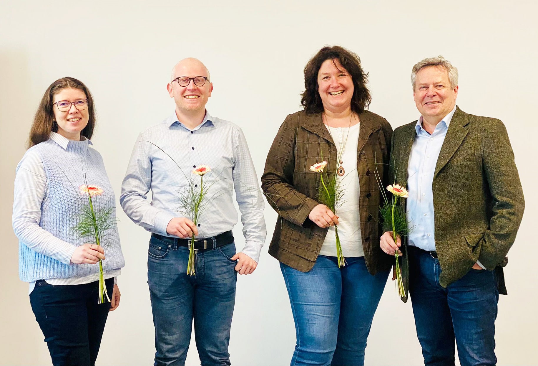 Vier Personen stehen nebeneinander und lächeln in die Kamera. Jede Person hält eine Blume in der Hand.