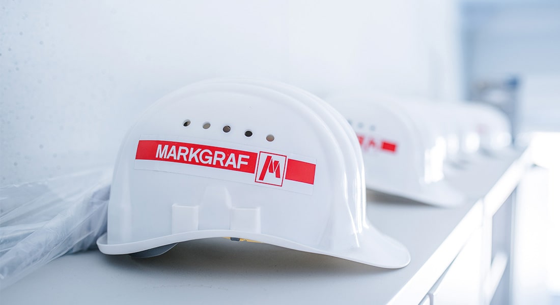 Ein weißer Arbeitshelm liegt neben anderen weißen Arbeitshelmen. Das Logo der Marke Markgraf ist auf dem vordersten Helm abgebildet.