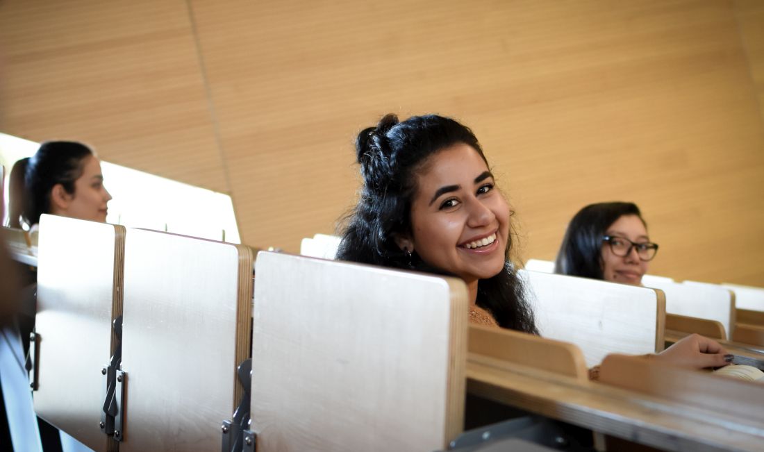Eine junge Frau mit schwarzen Haaren sitzt in einem Vorlesungssaal. Sie lächelt in die Kamera.