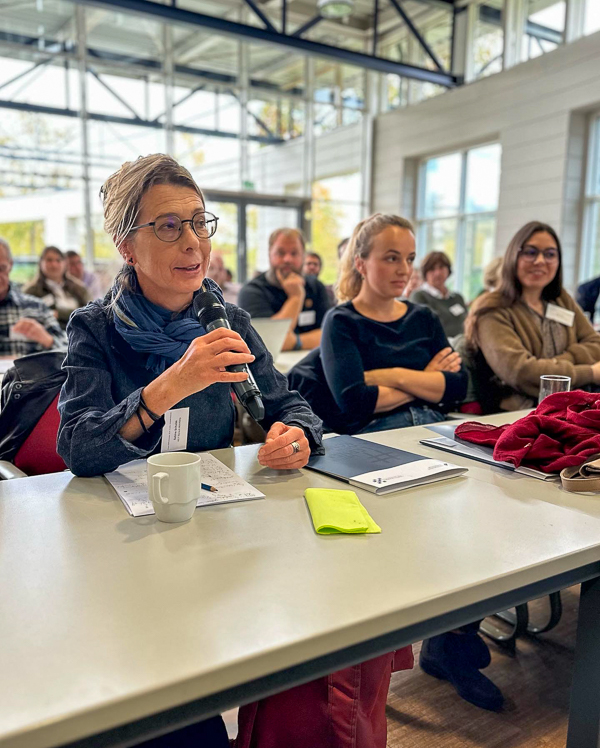 Weiblich gelesene Person sitzt in einem Publikum, redet und hält ein Mikrofon in der Hand.