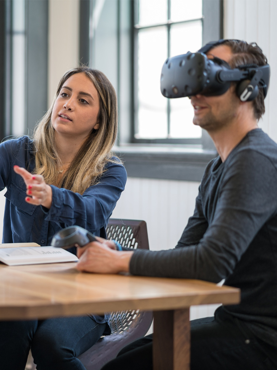 Zwei Personen in einem Büro, ein Mann mit VR-Brille und eine Frau, die in seine Blickrichtung gestikuliert