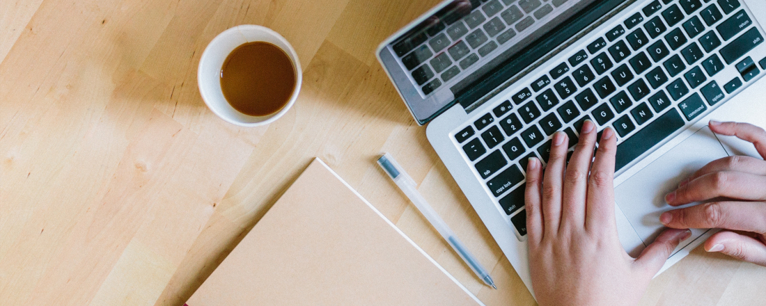 Hände bedienen Laptop auf einem Schreibtisch mit Kaffeetasse, Stift und Block daneben